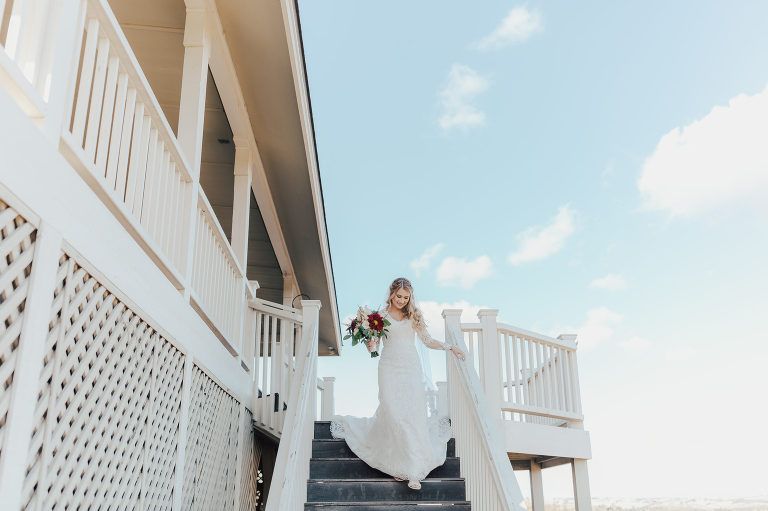 Bride in white dress descends outdoor stairs, holding bouquet, bright sky.