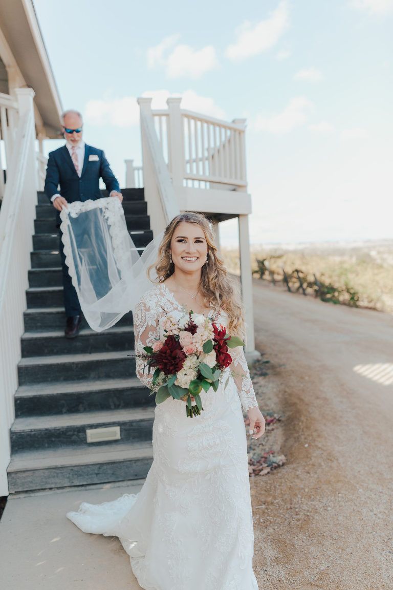 Bride smiles, descends stairs with veil, father follows behind. Outdoor wedding setting, bright light.