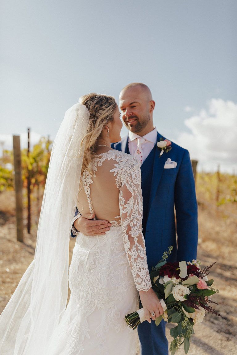 Bride and groom embrace outdoors; she in white lace gown and veil, he in blue suit; vineyard backdrop.