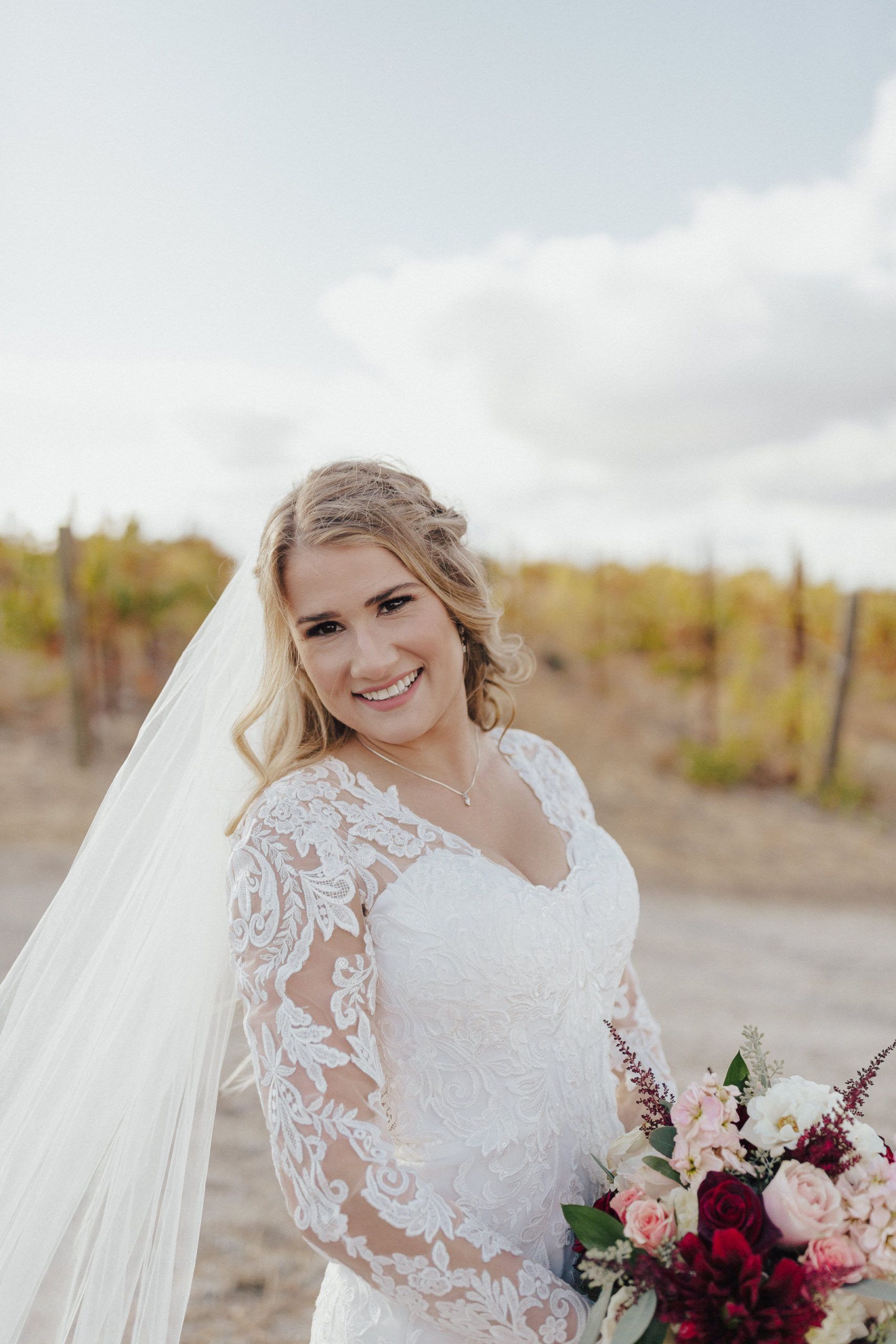 Bride in lace dress, smiling, holding bouquet, outdoors near vineyard.