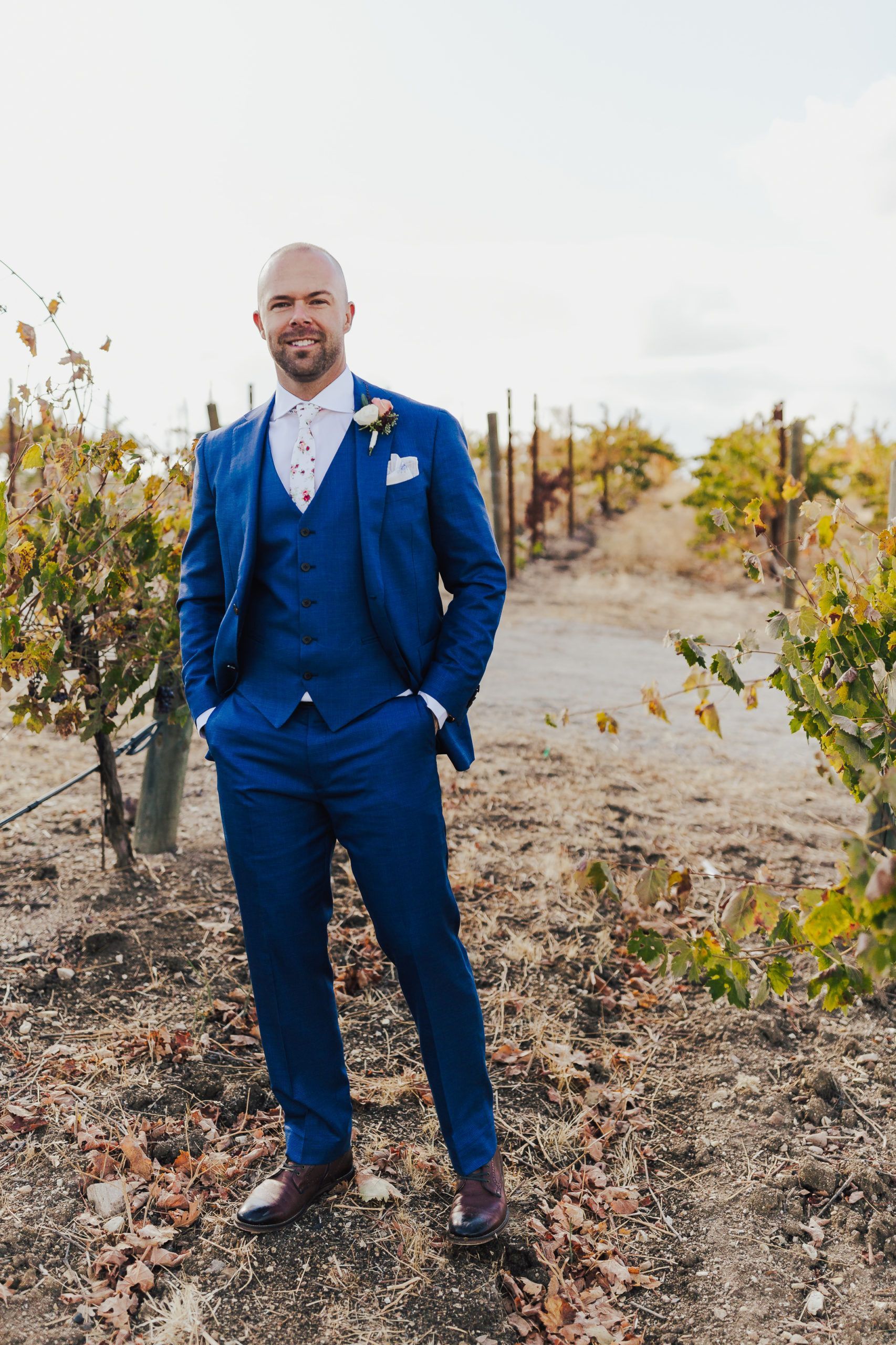 Man in blue suit stands in vineyard, smiling, hands in pockets.