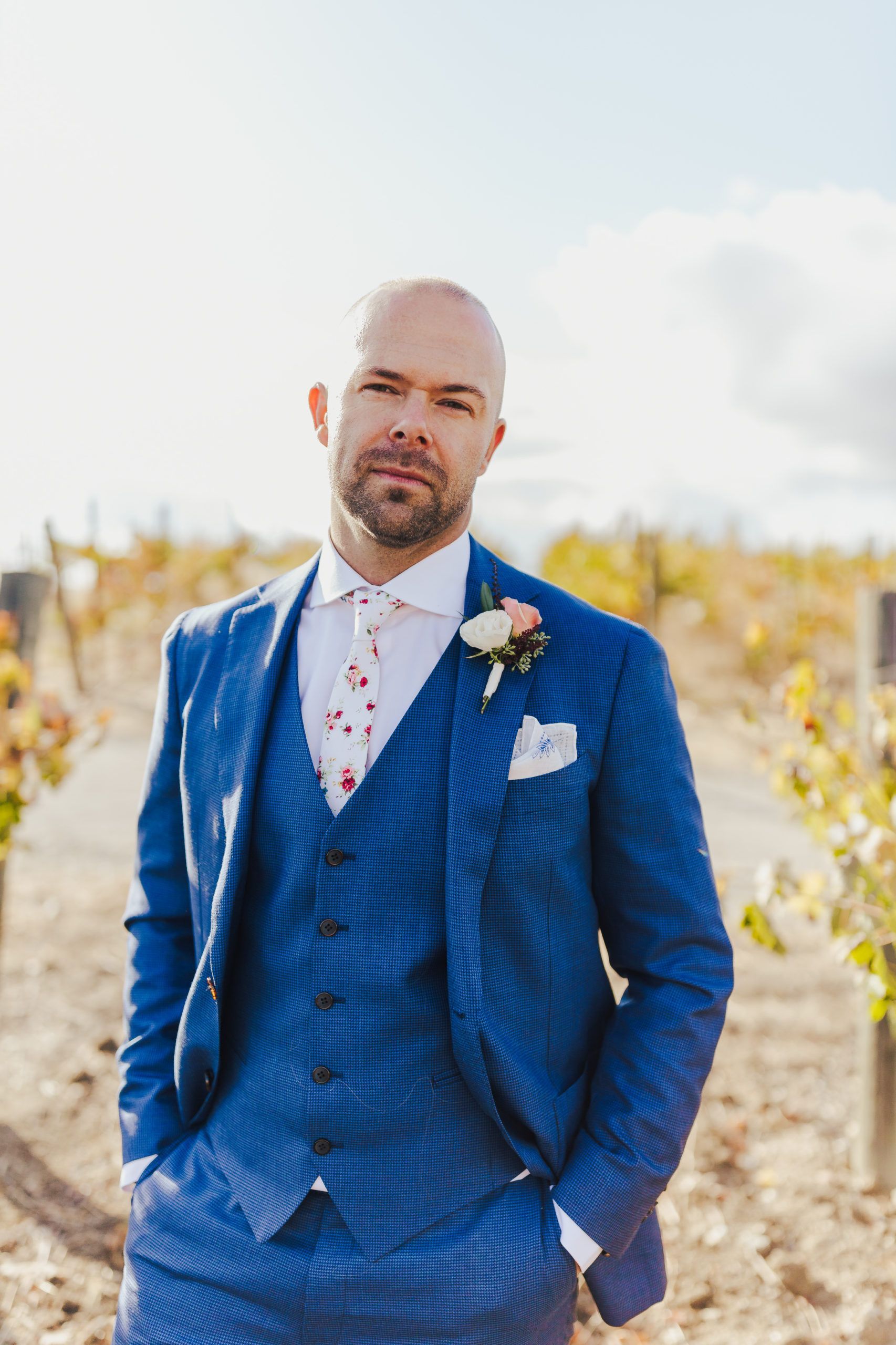 Man in blue suit, hands in pockets, stands in vineyard with a boutonniere and patterned tie.