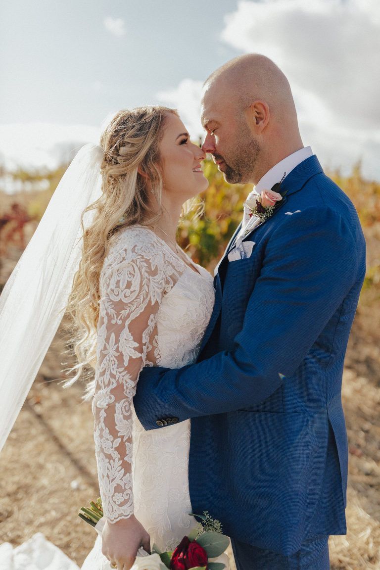 Bride and groom embrace outdoors, touching noses. She wears a lace dress, veil. He wears a blue suit.
