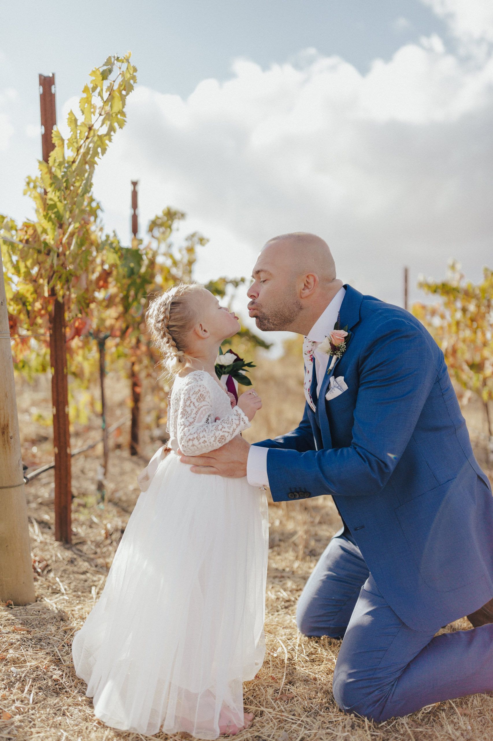 Man kneeling to kiss a child in a white dress at a vineyard.