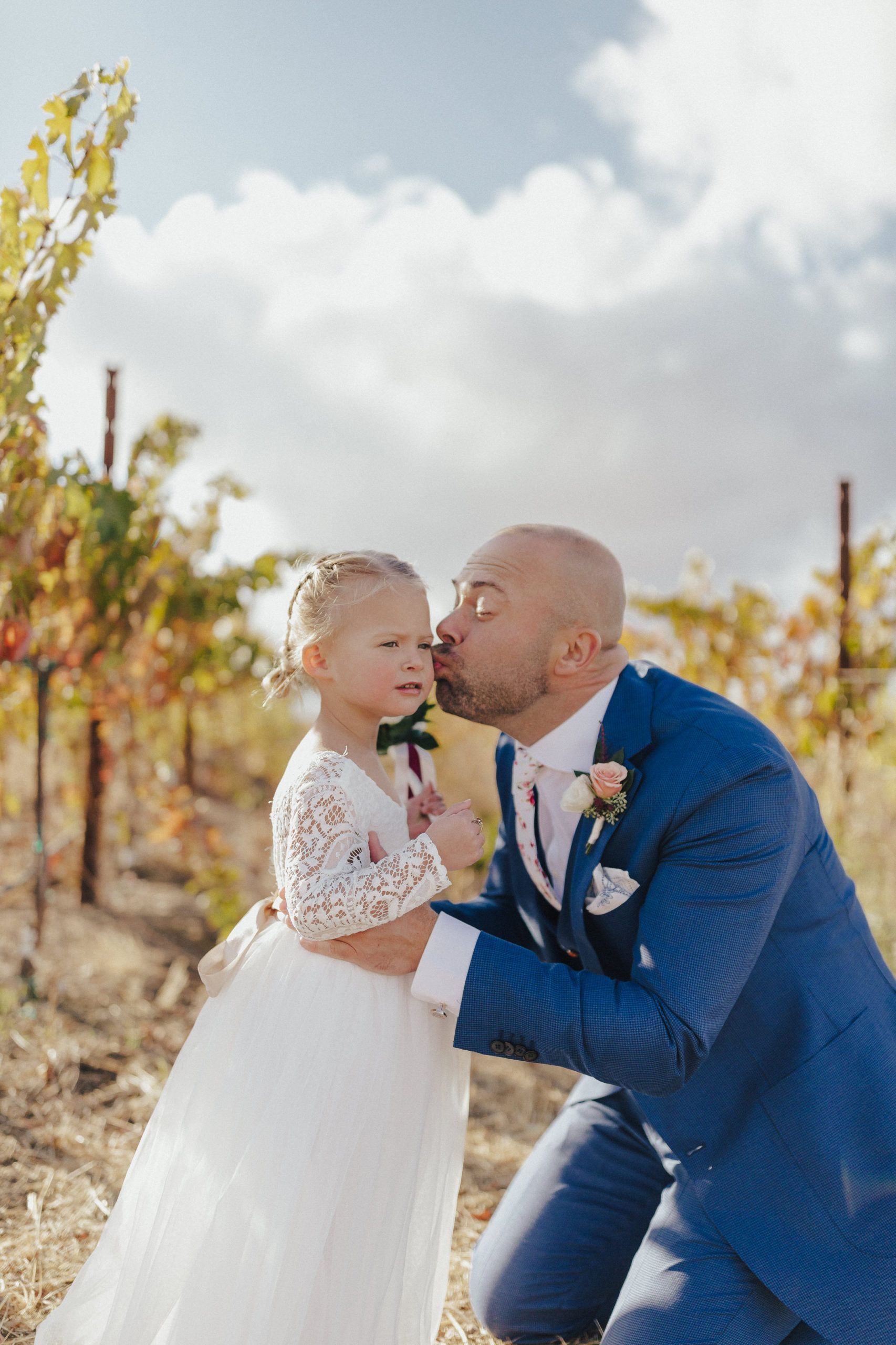 Man in blue suit kisses a young girl in white dress on the cheek in a vineyard.