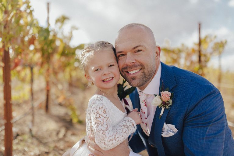 Man in blue suit embraces girl in white dress; vineyard background. Both smiling.