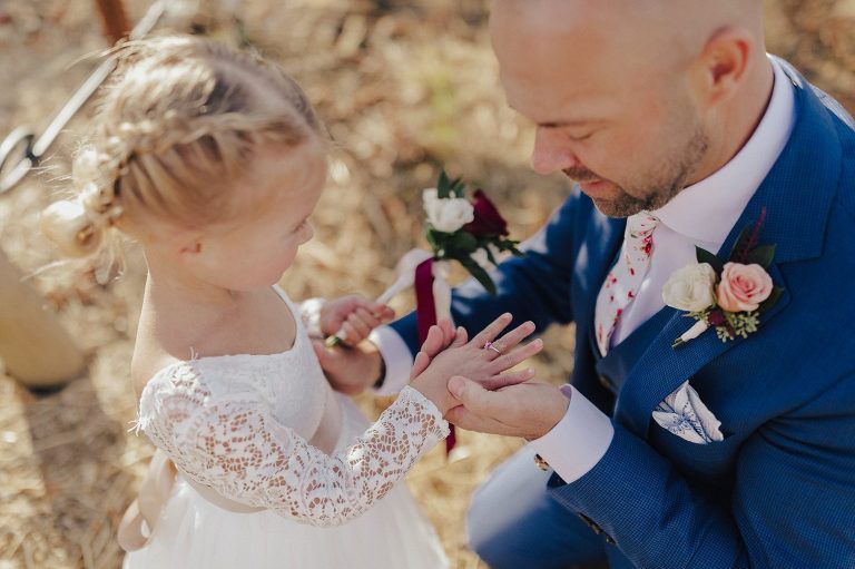 Man and flower girl examining hand with wedding rings, outdoors. Man in blue suit, flower girl in white dress.