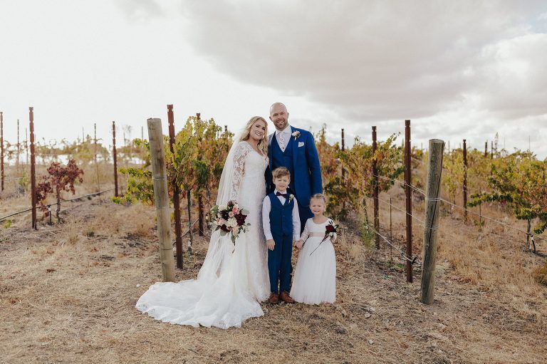 Family poses for a photo at a vineyard wedding. The couple and two children are smiling in front of grapevines.