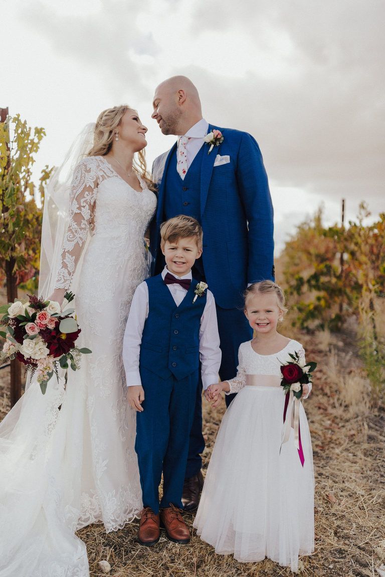 Wedding group in vineyard: Bride in lace dress, groom in blue suit, holding hands with two children.