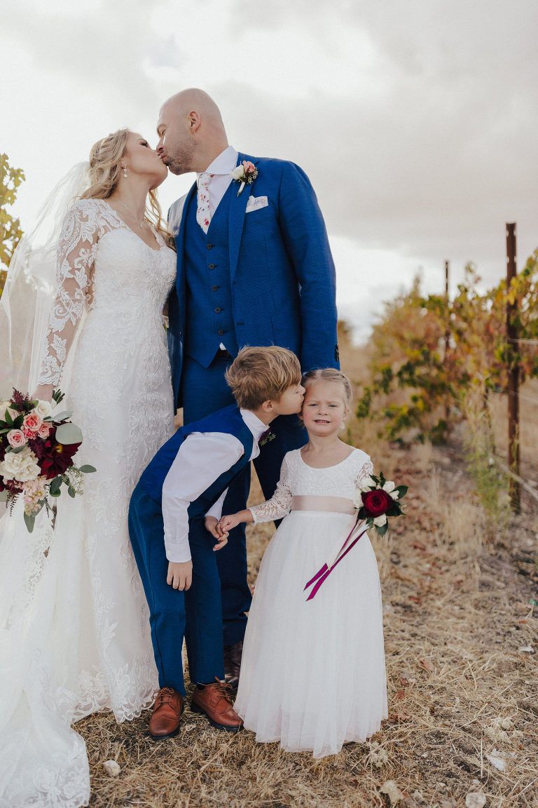 Newlyweds kissing, flanked by children, in a vineyard. Bride in lace dress, groom in blue suit. Autumn colors.