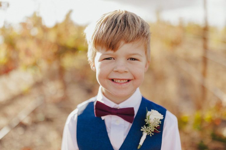 Boy in a blue vest and burgundy bow tie smiles, a small flower on his lapel; outdoors with blurred background.