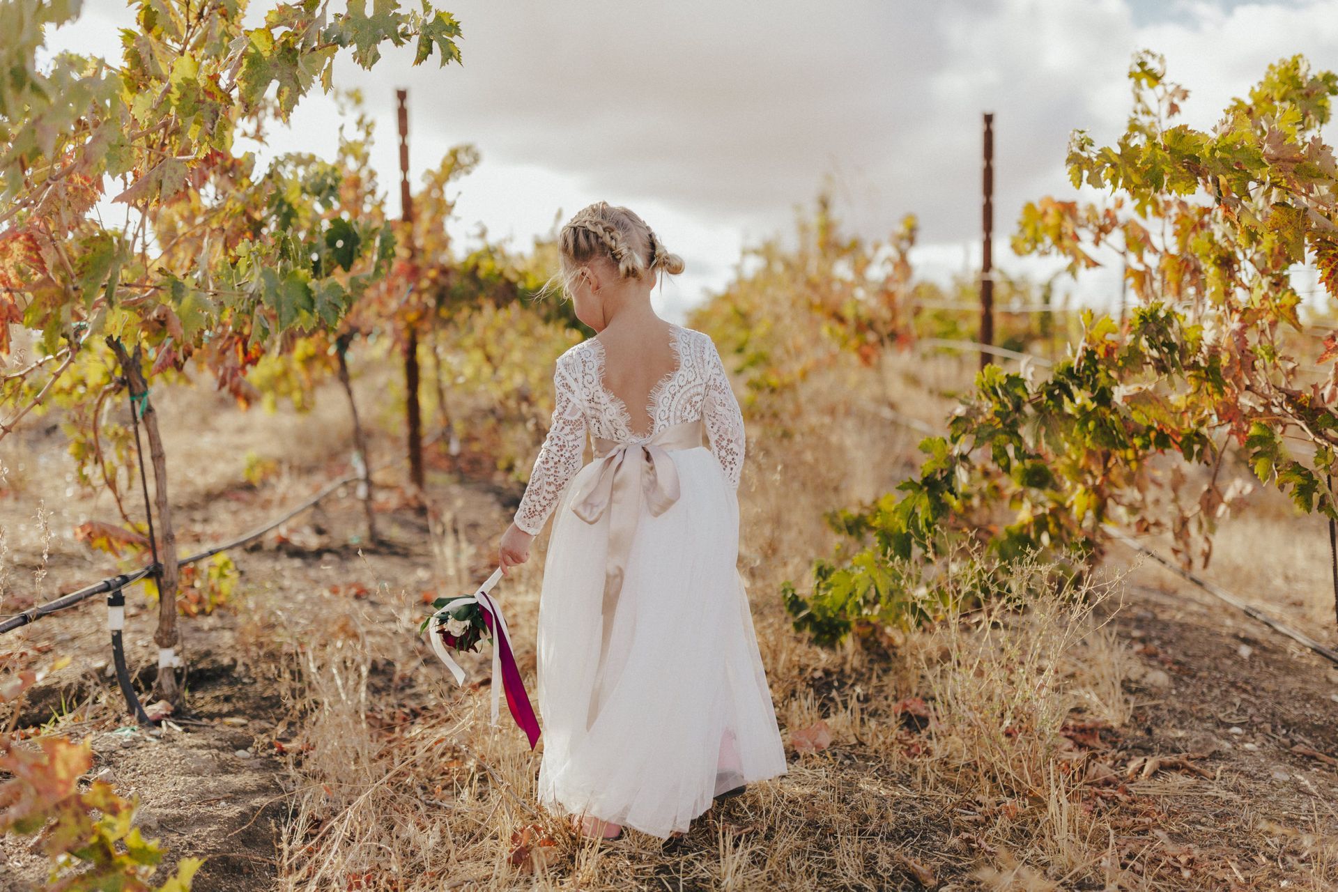 A young girl in a white dress stands in a vineyard, holding a bouquet, with fall foliage in the background.