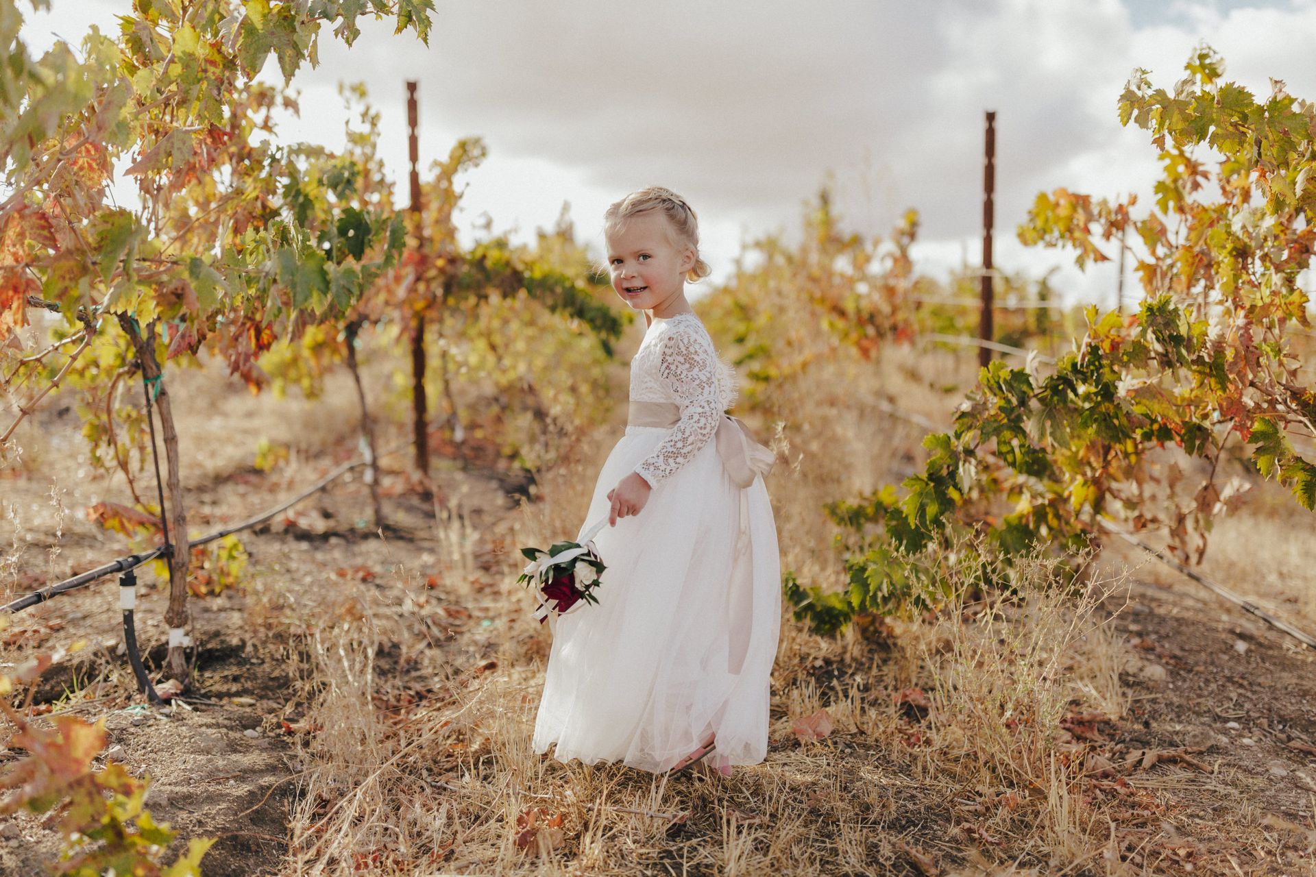 Young girl in white dress smiles, holding flowers, standing in a vineyard.