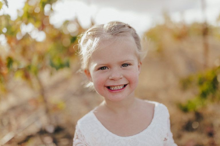 Blonde girl smiling, wearing a white dress, outdoors with blurred yellow background.