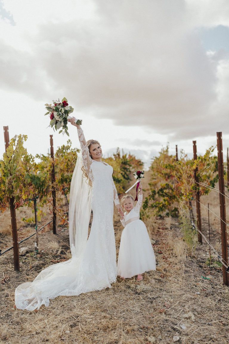 Bride holding bouquet with flower girl, in a vineyard. Both are raising arms in celebration.