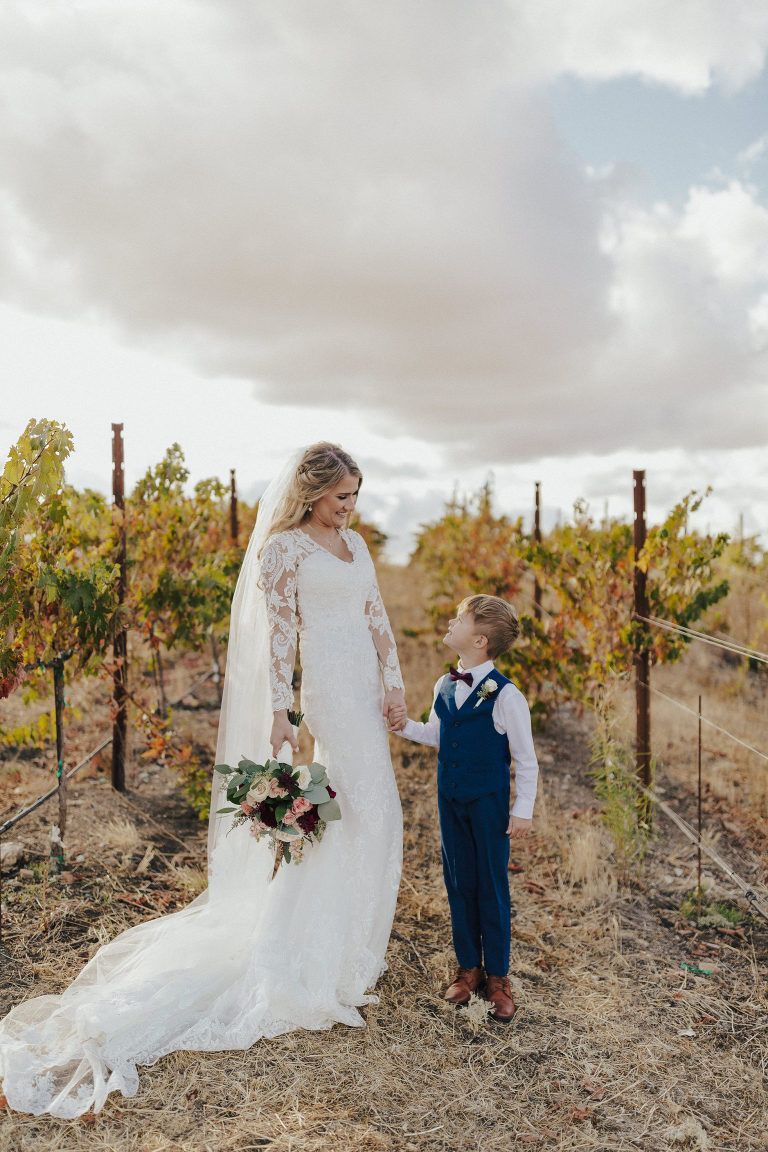 Bride in lace dress holds hands with a young boy in a blue suit in a vineyard.