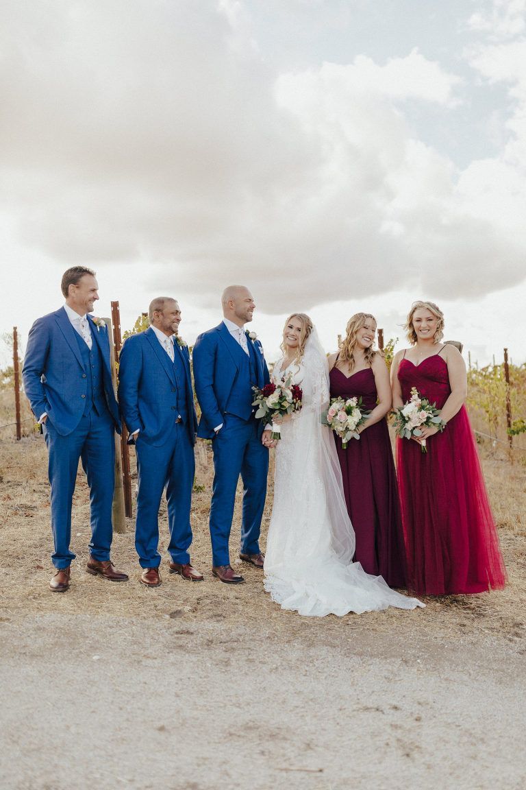 Wedding party poses outside. Groomsmen in blue suits, bridesmaids in burgundy dresses. Bride wears white. Sunny day.