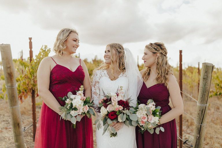 Bride smiles with two bridesmaids in a vineyard, all holding bouquets.