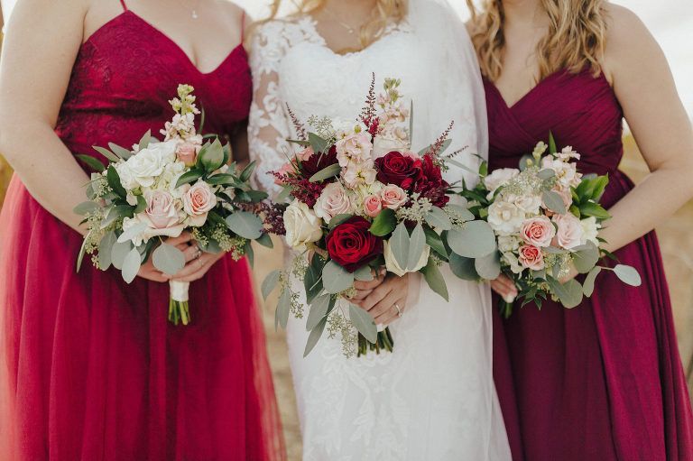 Bride with bridesmaids holding bouquets, wearing burgundy gowns, outdoors.