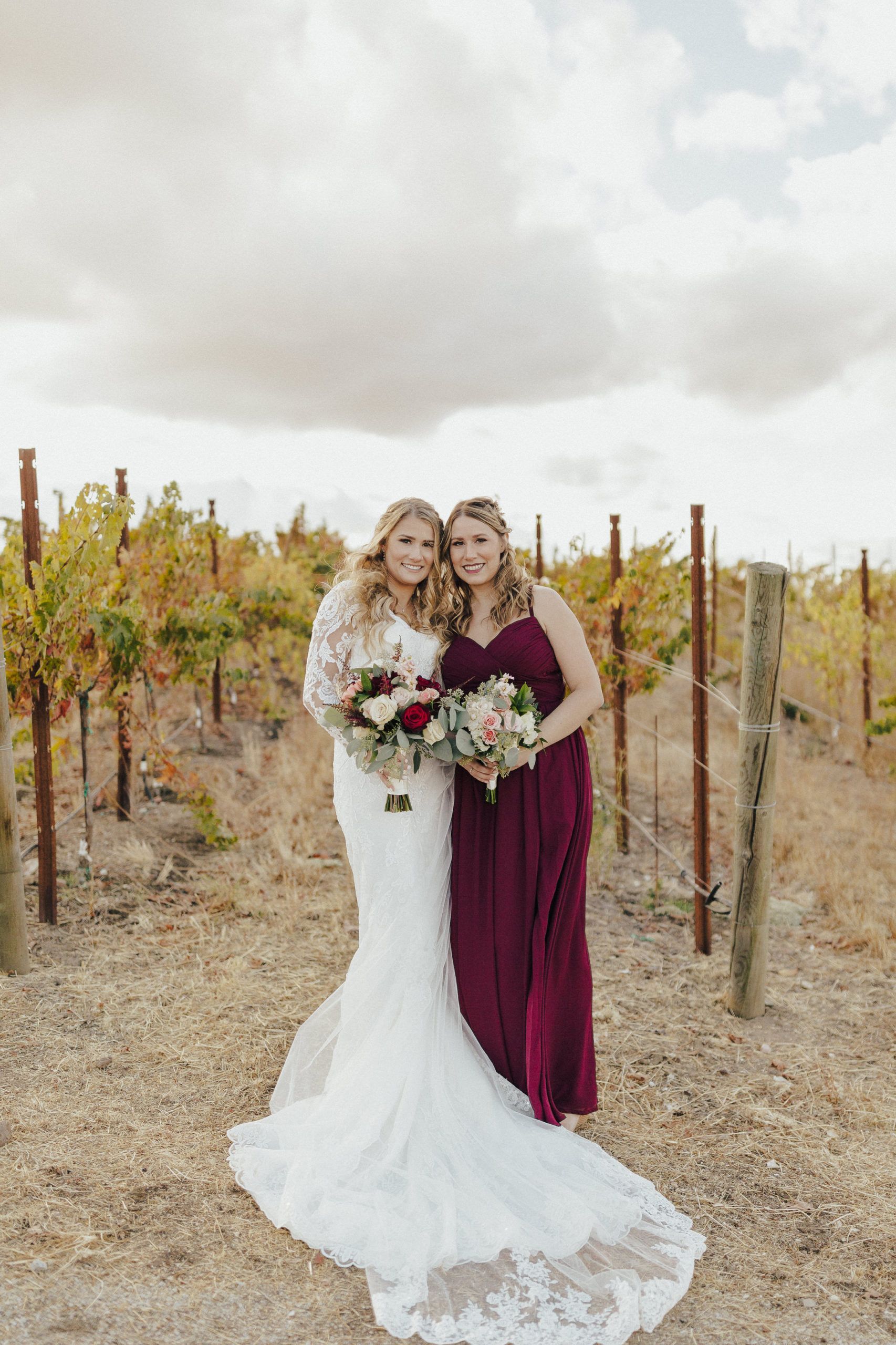 Bride in lace dress and bridesmaid in burgundy dress holding bouquets in vineyard.