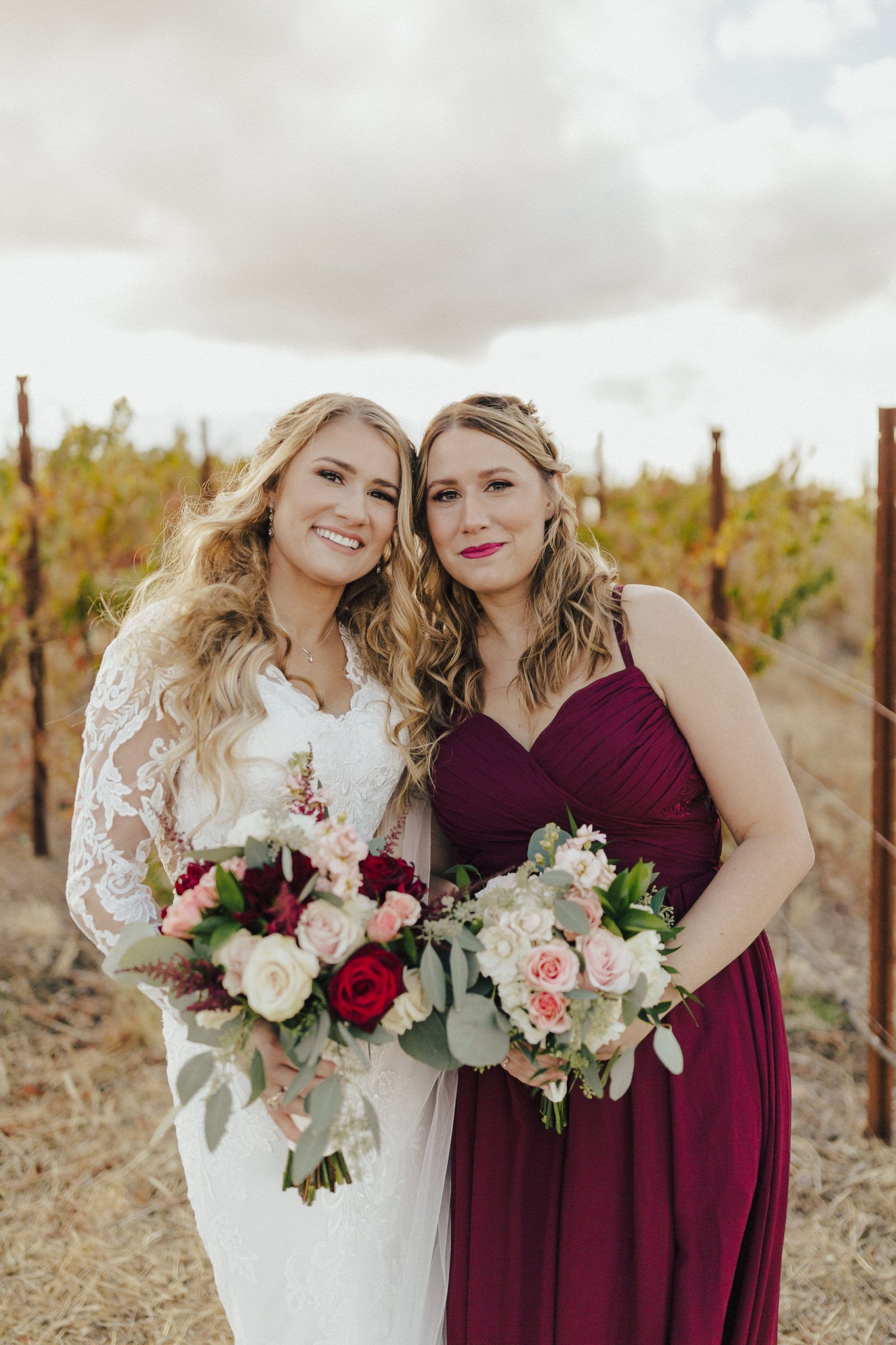 Bride and bridesmaid pose in vineyard, holding bouquets. The bride wears white lace, bridesmaid in burgundy.