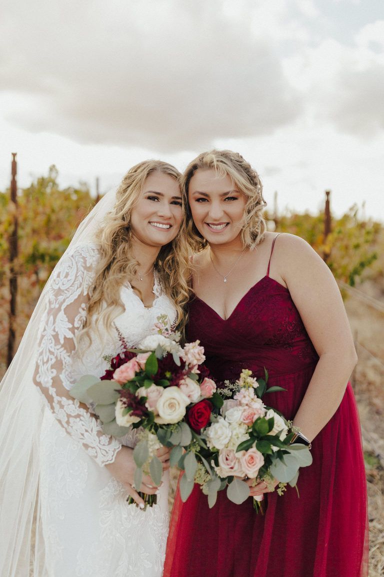 Bride in lace dress and bridesmaid in burgundy gown with bouquets, smiling in vineyard.