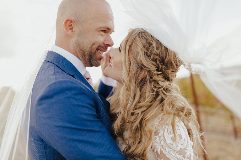 Wedding couple embracing, bride's hair braided. Man in blue suit, woman in lace dress, veil blowing in the wind.