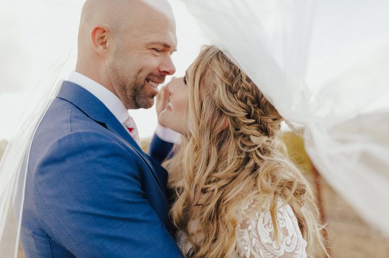 Bride and groom face each other, under flowing veil. She has long blonde hair; he wears a blue suit.