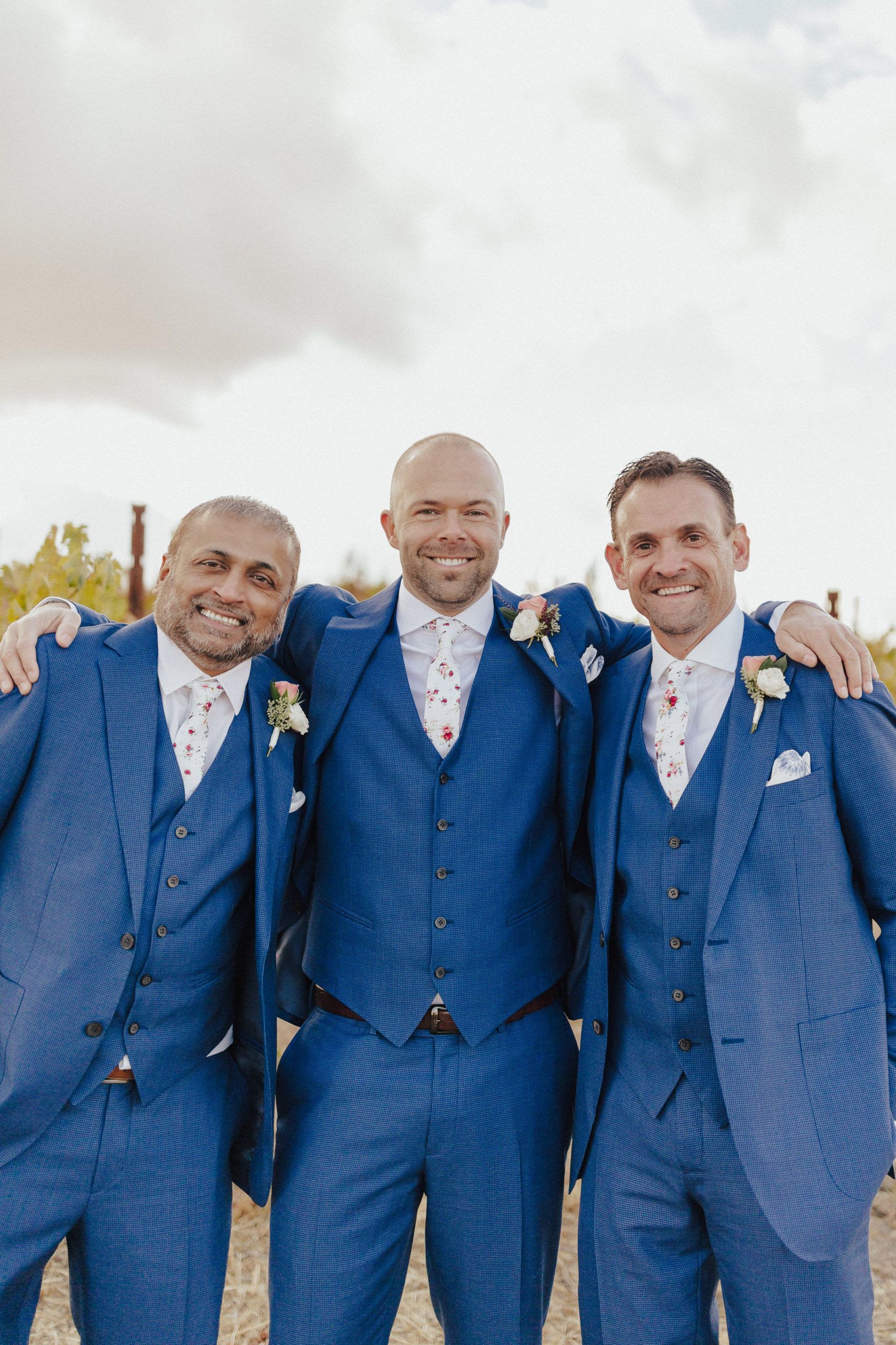 Three men in blue suits stand close, arms around each other, smiling, outdoors.