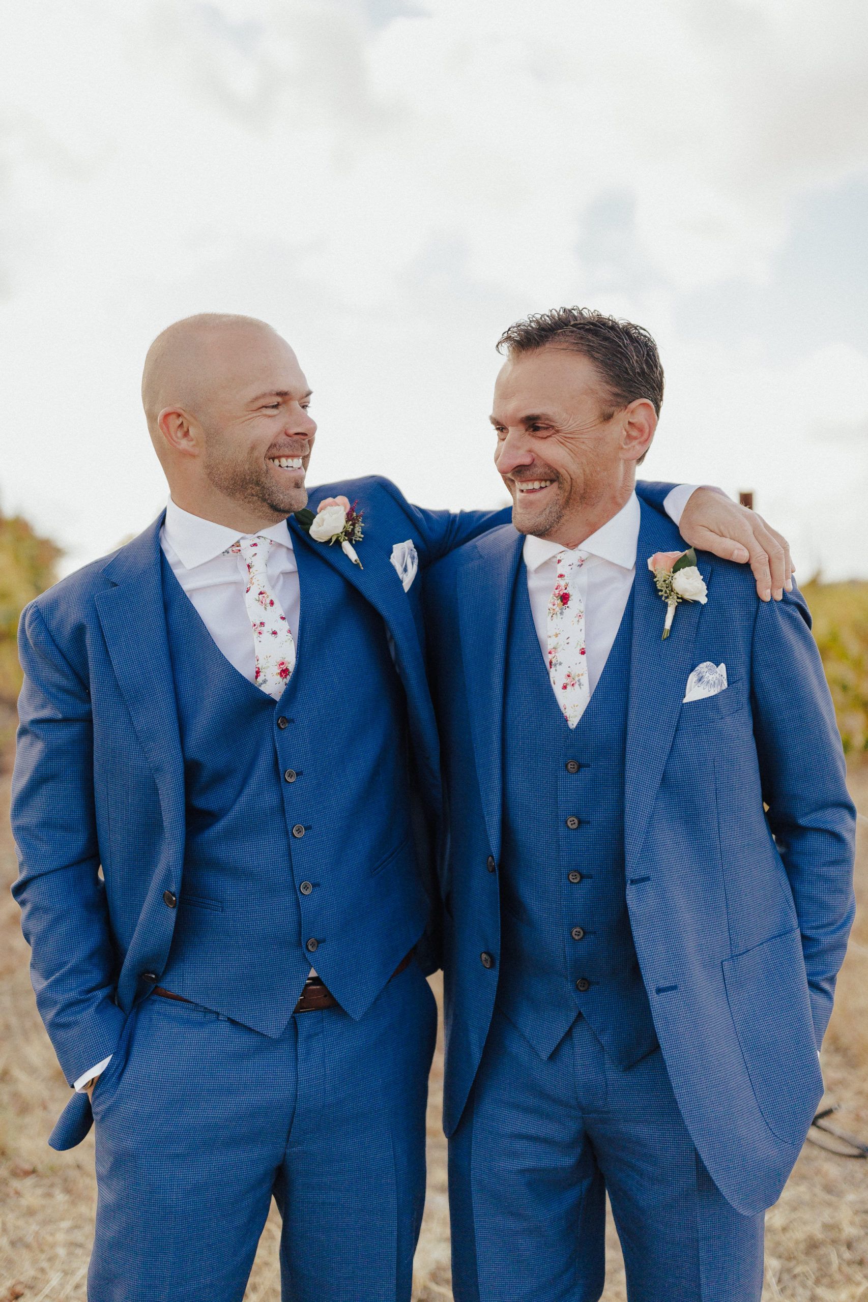 Two men in blue suits smile, embracing outdoors.