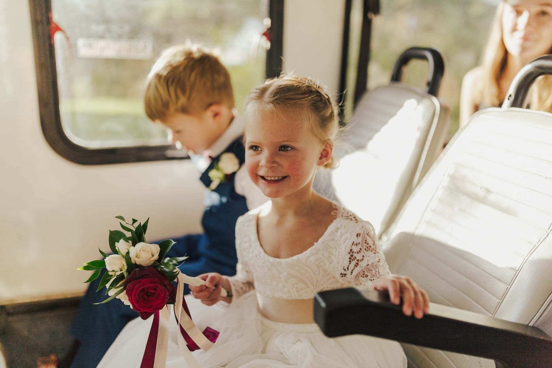 Flower girl smiling in a white dress, holding a bouquet, sits in a vintage vehicle with a boy in a blue suit.