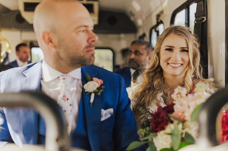 Bride and groom in wedding attire on a bus, smiling. Groom in blue suit, bride holding bouquet.