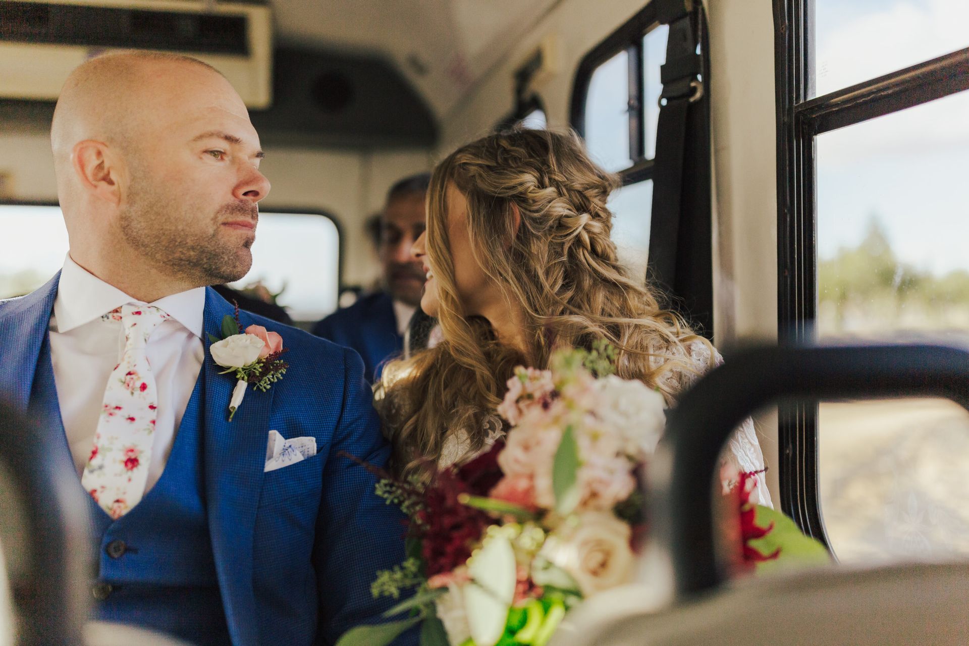 Bride and groom on a bus, the man in a blue suit looks to the left, woman holds a bouquet.
