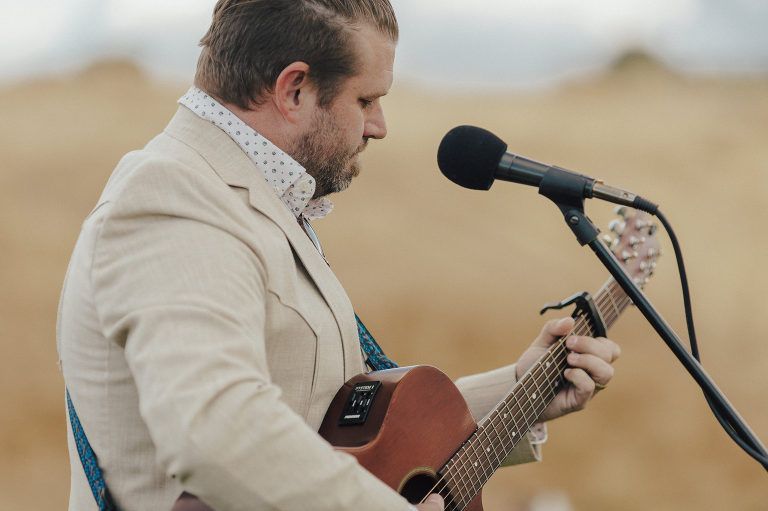 Man playing acoustic guitar, singing into a microphone. He wears a light-colored suit jacket, outdoors in a field.