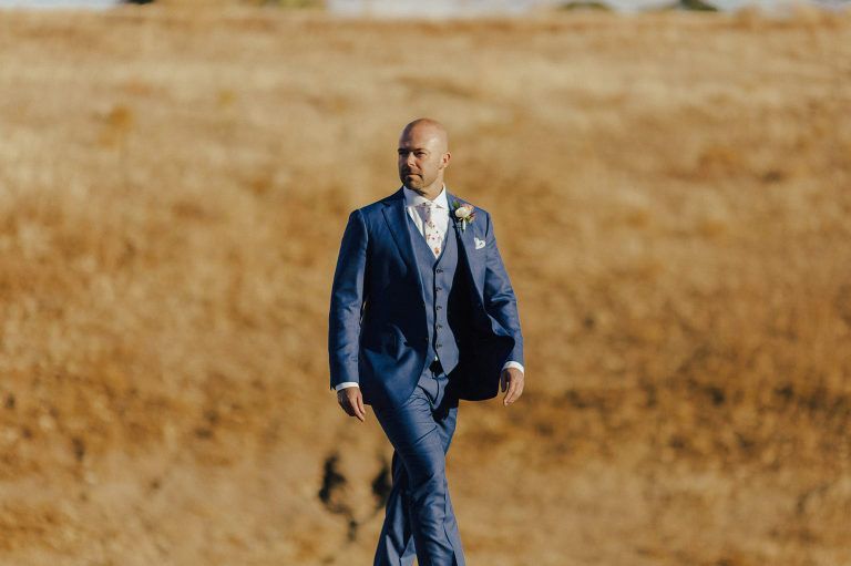 Man in blue suit walks across a field, wearing a boutonniere.