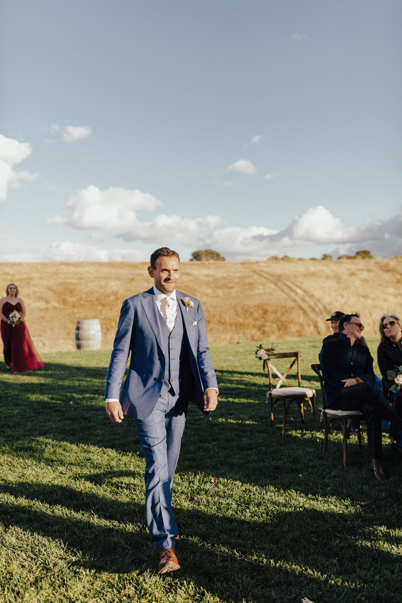 Man in blue suit walks down grass aisle towards camera, wedding ceremony. Sunny field.