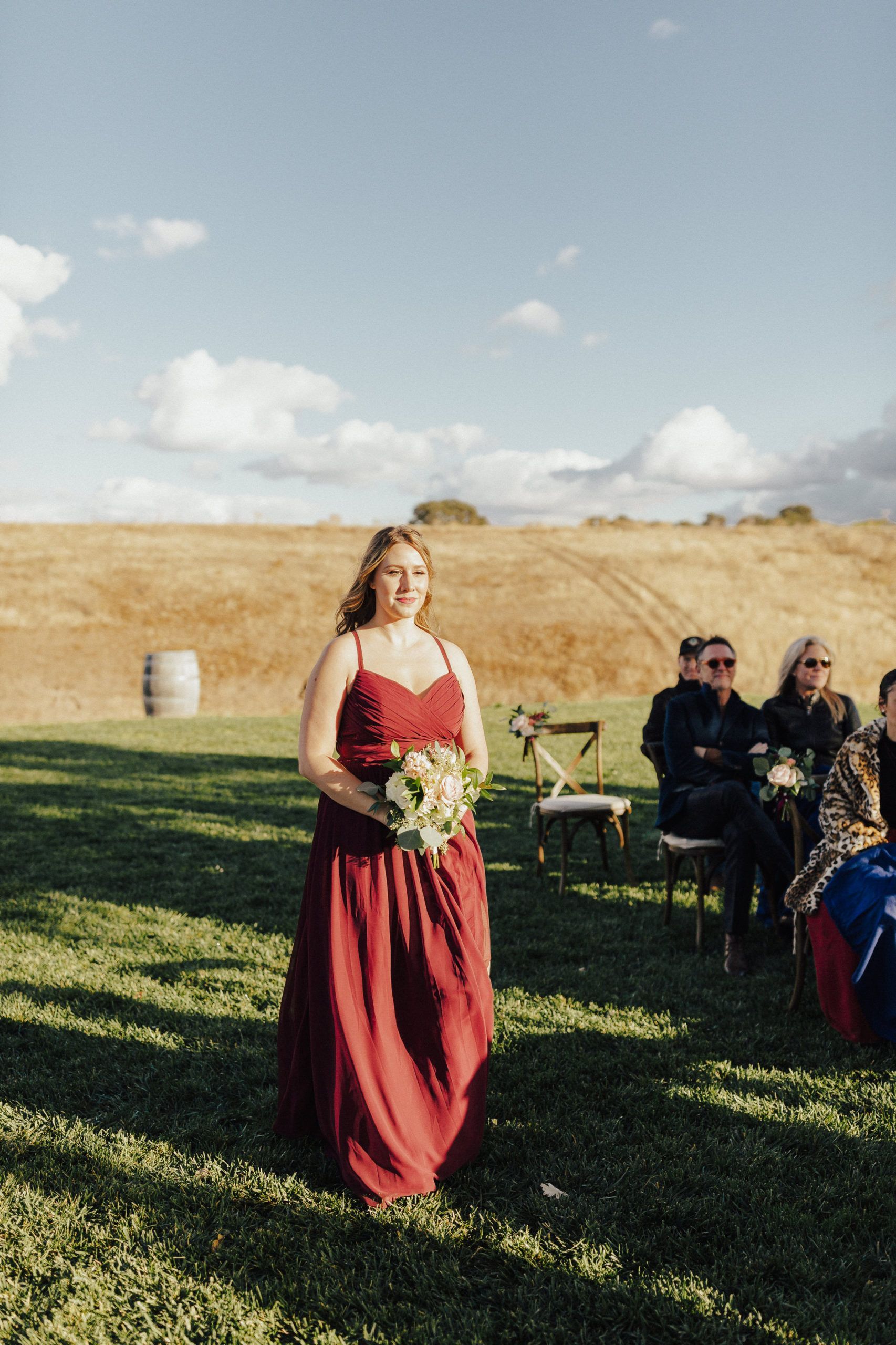 Woman in a red dress carrying flowers stands outside on a sunny day at a wedding ceremony.