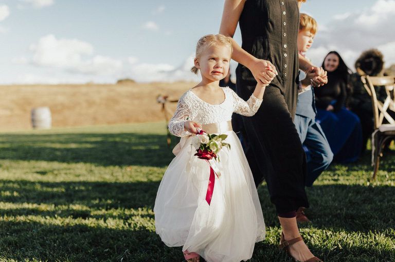 Flower girl in white dress and bouquet walks holding hands. Other child and adult in background on green grass.