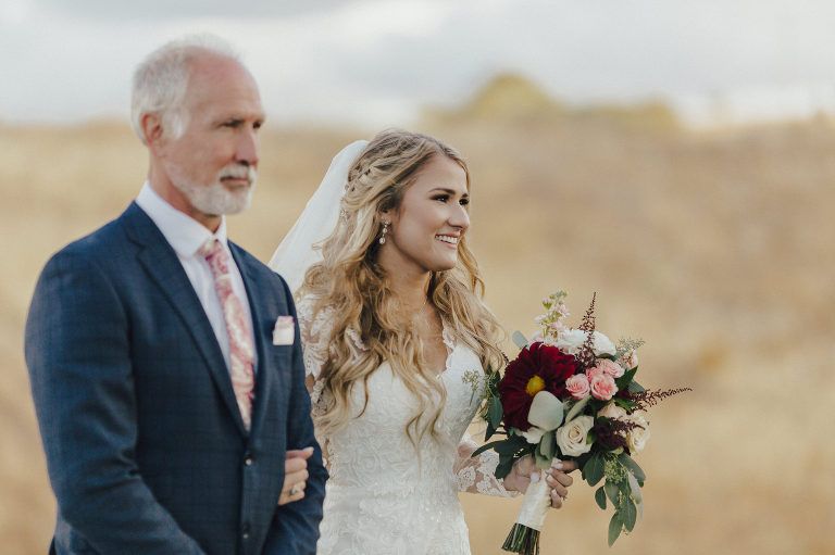 Bride and her father walking outdoors; bride in white dress, holding bouquet; father in navy suit.