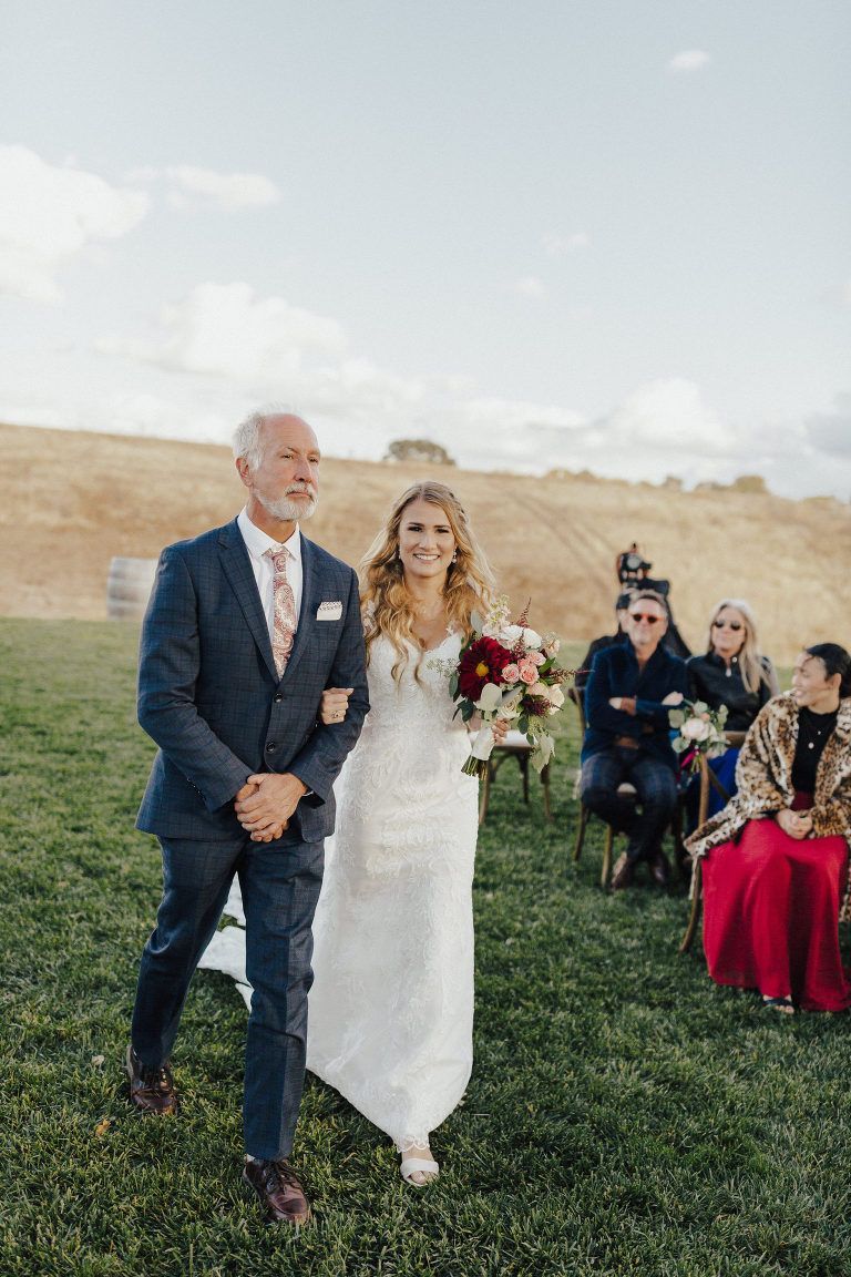 Father walks bride down aisle at outdoor wedding. Green grass, rolling hills, guests seated.