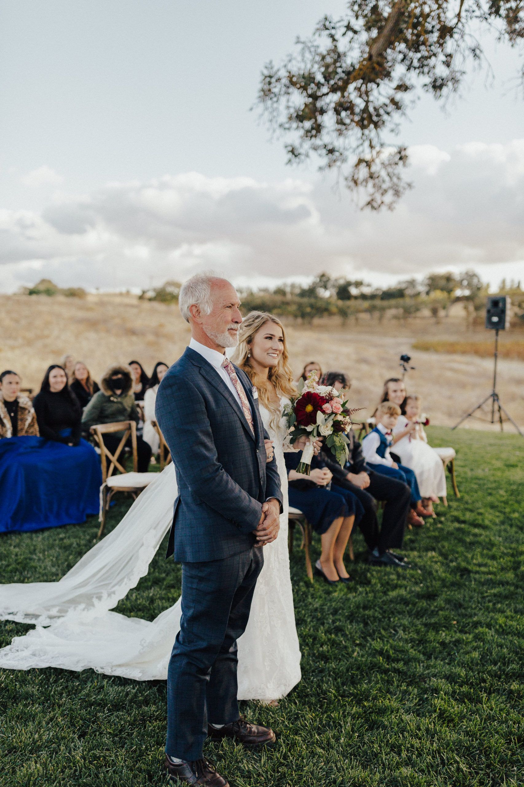 Bride and father at outdoor wedding ceremony. Bride in white dress, father in navy suit, smiling, watching the ceremony.