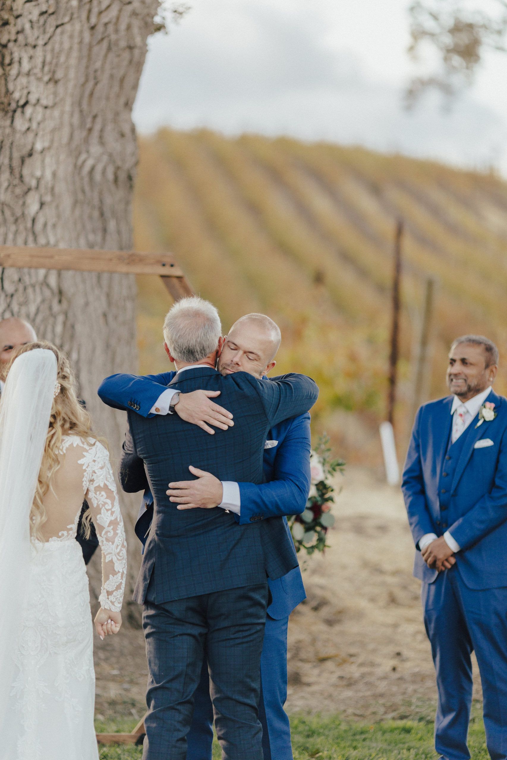 Two men embracing at an outdoor wedding ceremony; one in a blue suit, the other in a dark suit, vineyard backdrop.