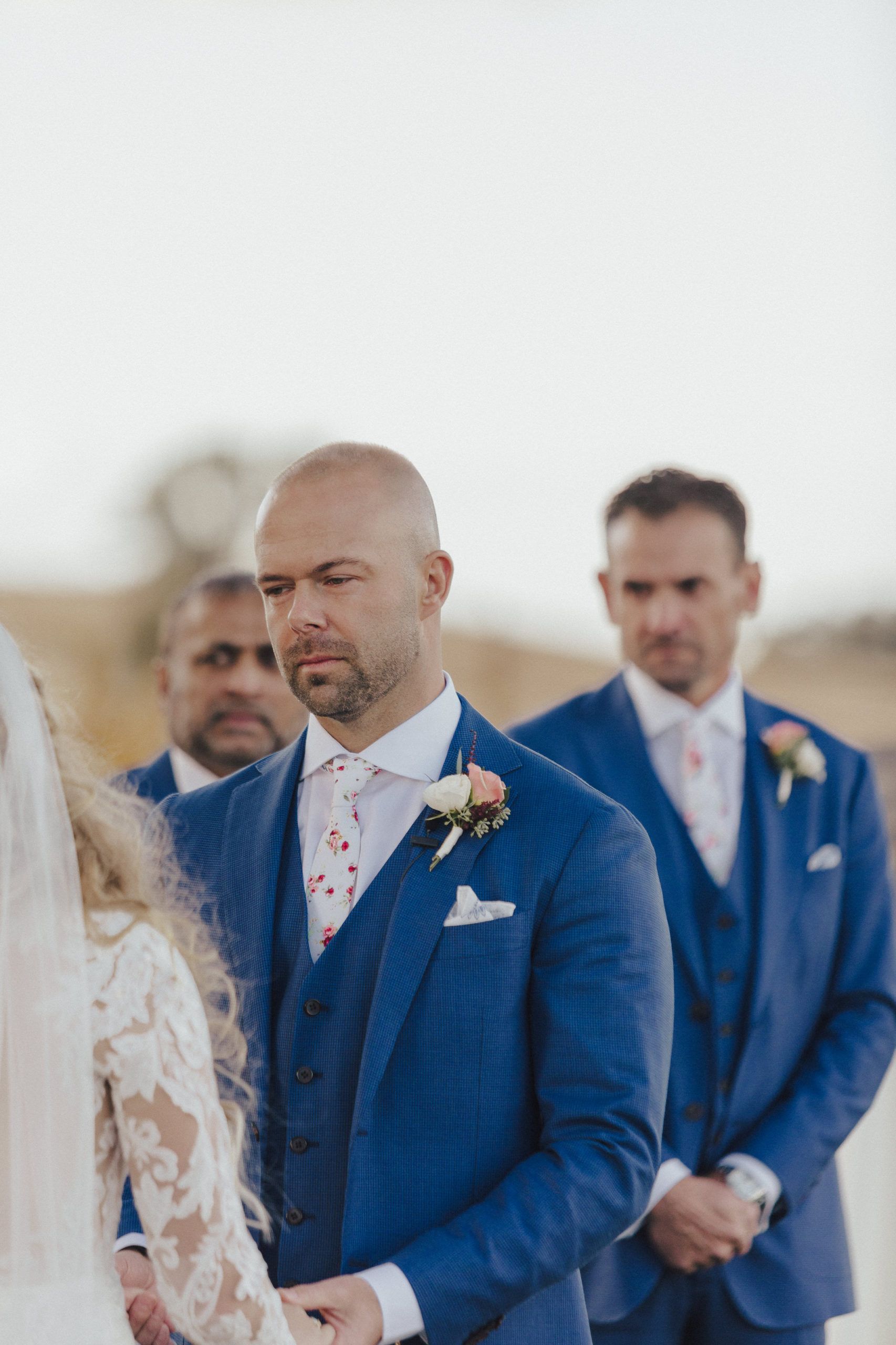 Groom in blue suit at outdoor wedding ceremony; he looks emotional.