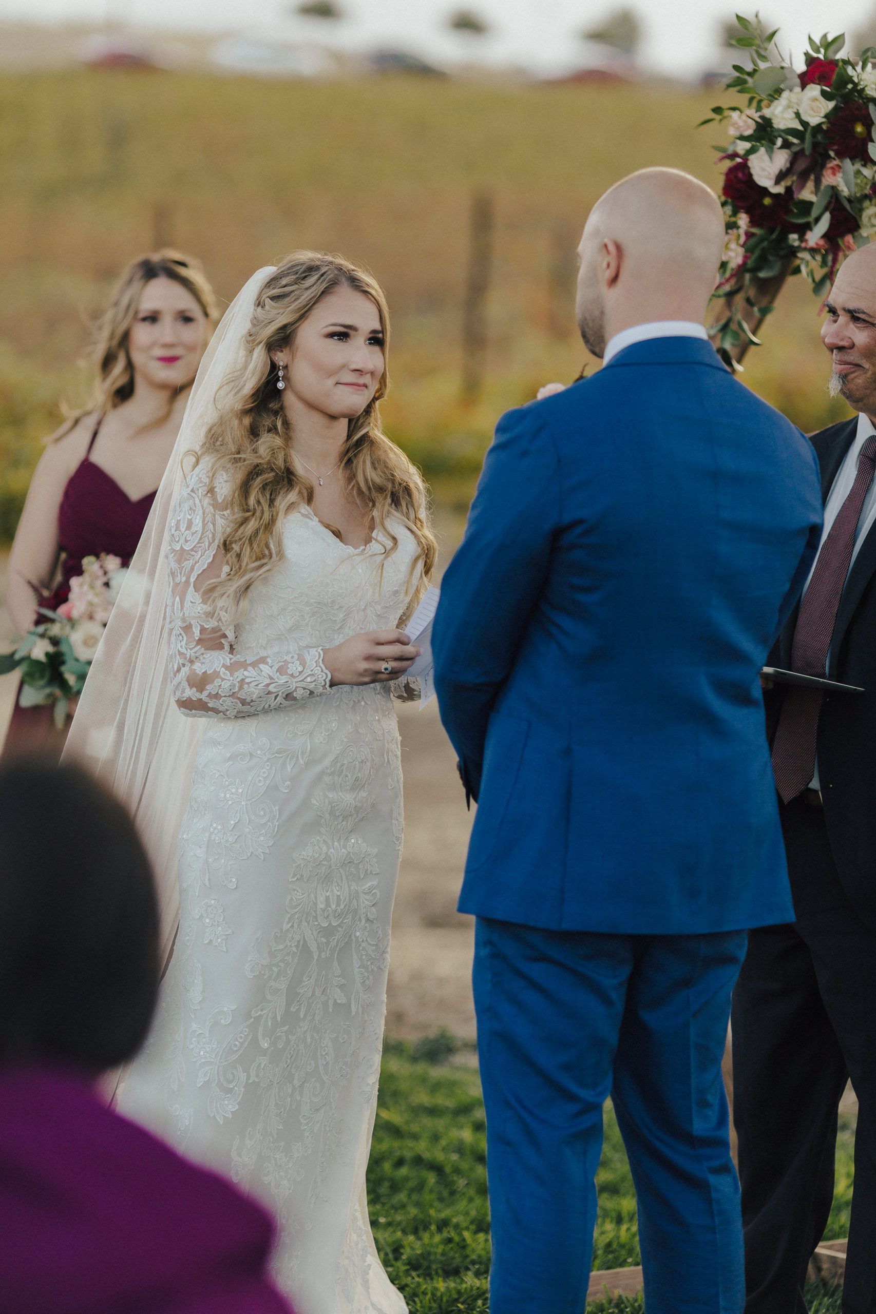 Bride in lace gown looks at groom in blue suit at outdoor wedding.
