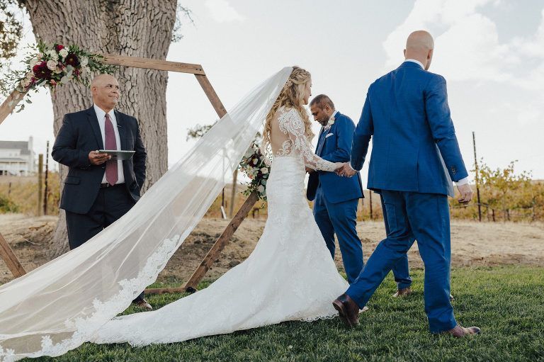 Bride walking down the aisle, holding hands with groom, toward ceremony arch. Veil and dress train trailing.