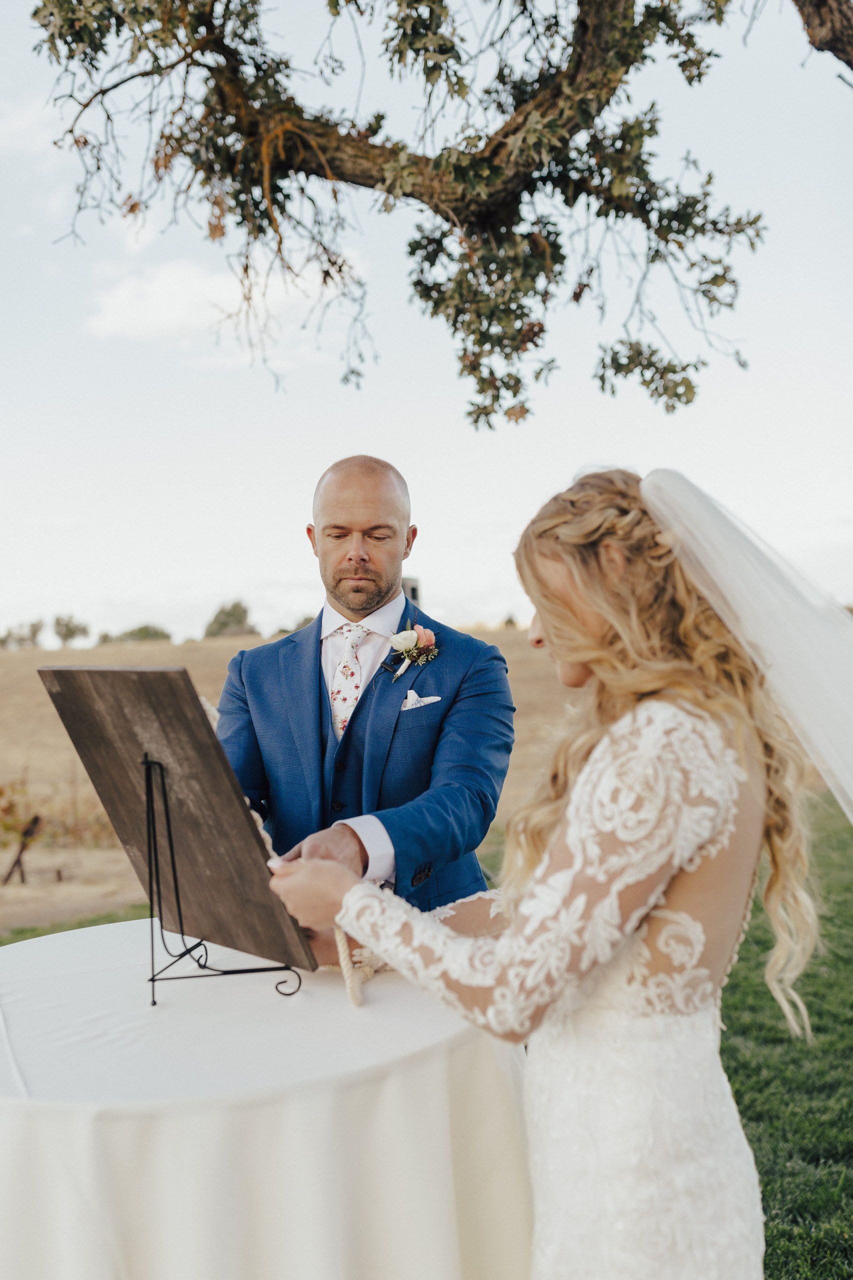 Bride and groom signing a wooden guest book at outdoor wedding; groom in blue suit, bride in white dress.