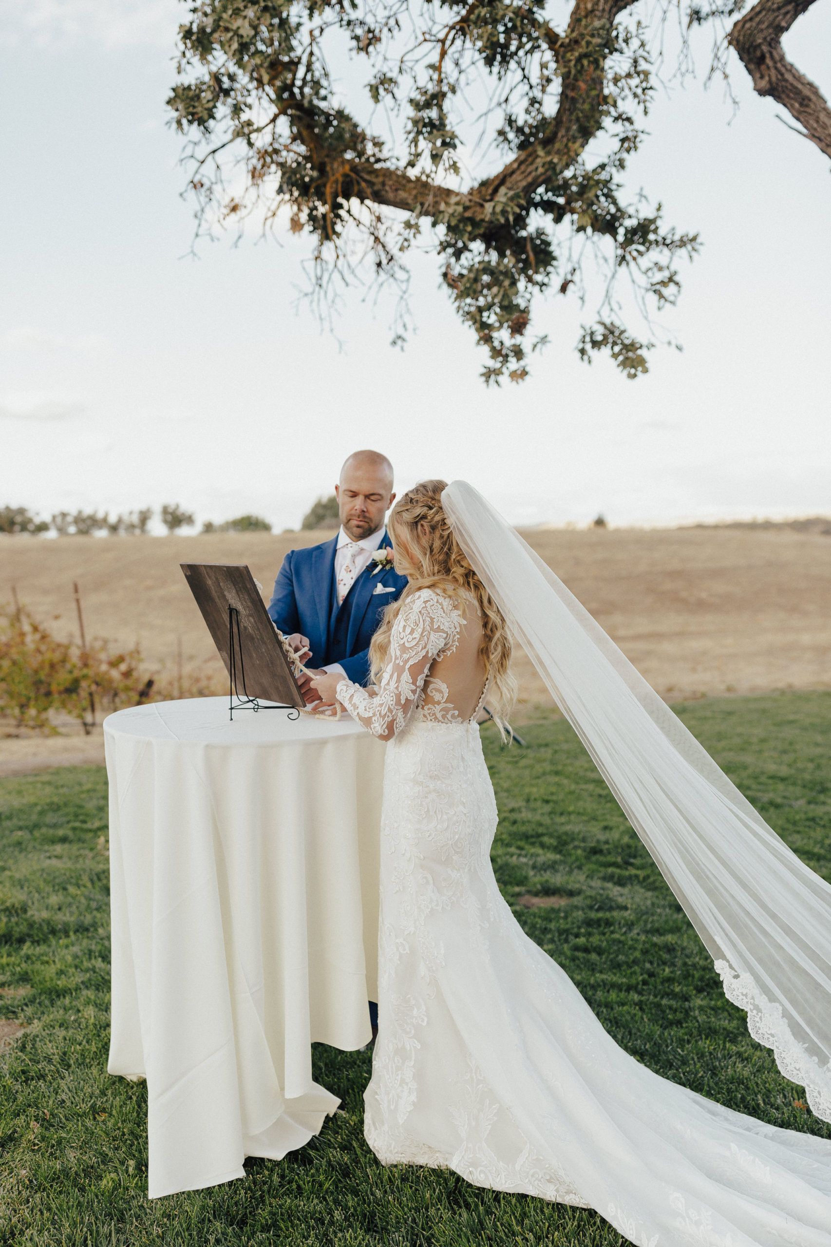 Bride and groom signing marriage documents at outdoor ceremony. Bride in white gown with veil, groom in blue suit.