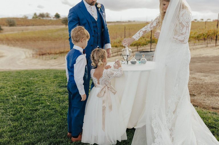 Wedding sand ceremony with family in a vineyard. Bride, groom, and children pour sand into a vase.