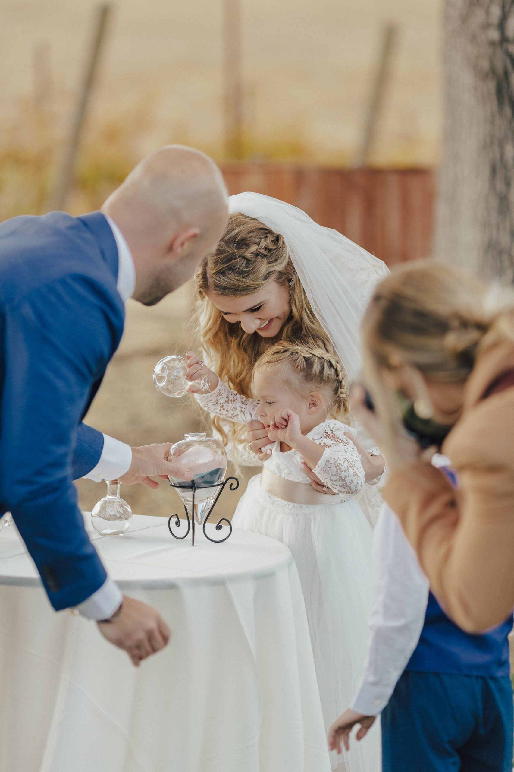 Bride, groom, and flower girl at a wedding. Pouring into a unity candle holder on a table outdoors.