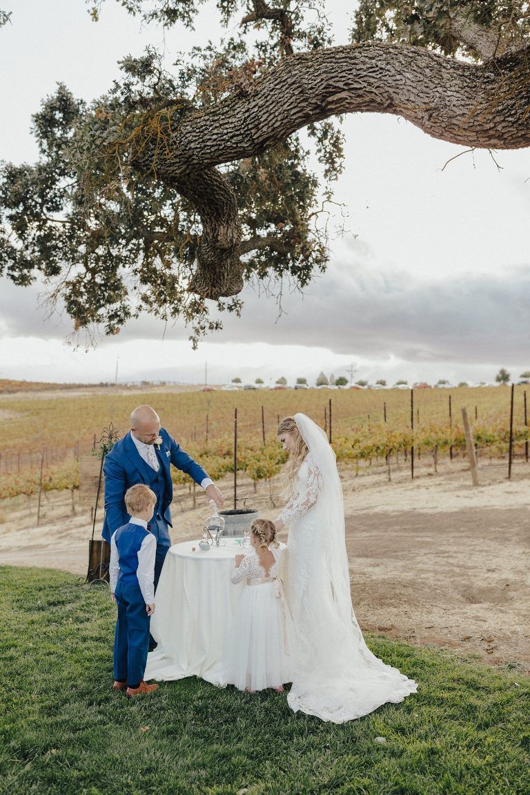 Family at outdoor wedding ceremony; pouring sand, bride in white gown, blue-suited groom, vineyard backdrop.
